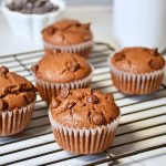 Whole wheat double chocolate chip muffins placed on wire rack with a mug of coffee in the back