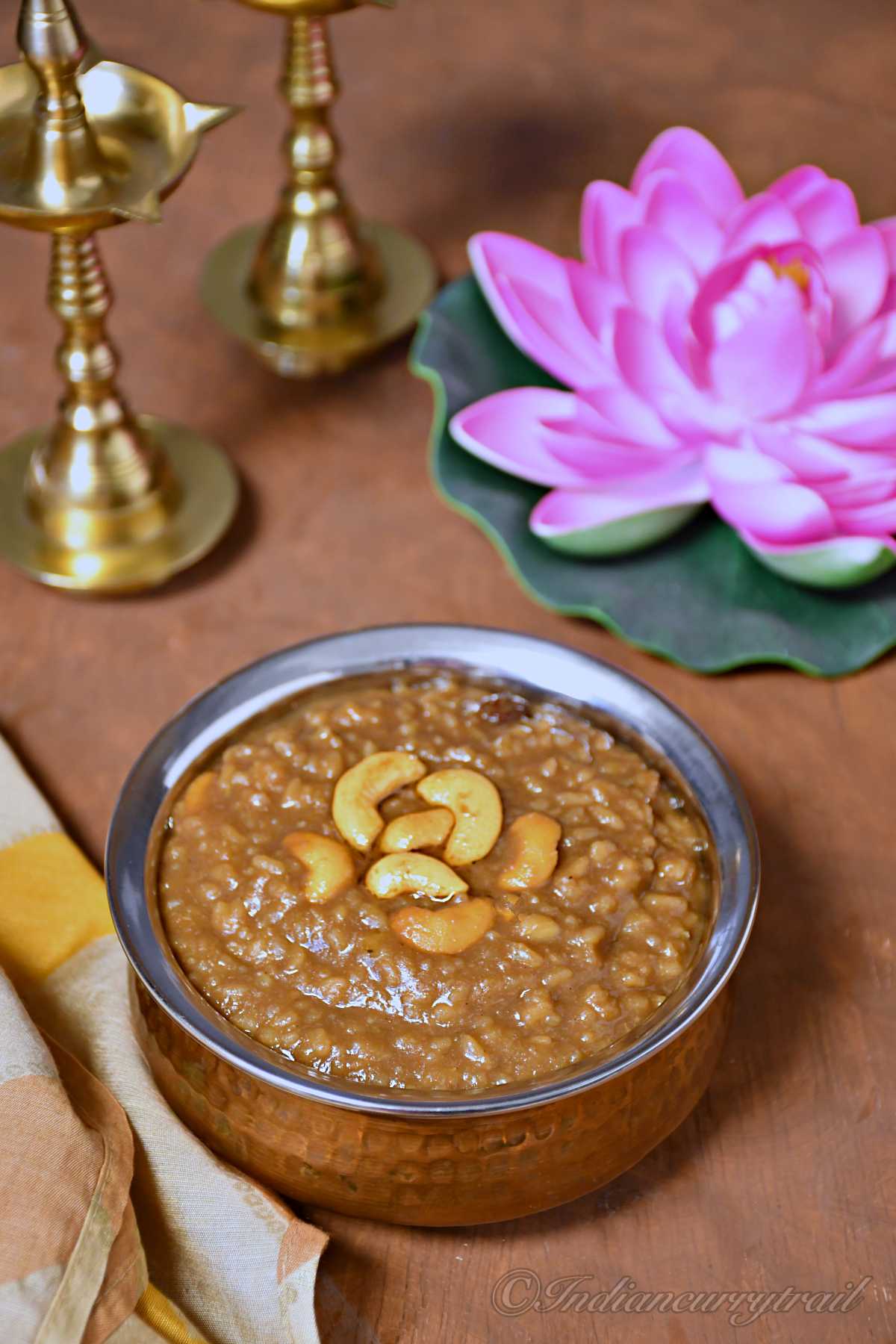 a picture showing a bowl of palm jaggery pongal with fried cashew nuts on top of it and traditional indian diya and lotus behind