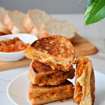 a stack of kimichi grilled cheese sandwich kept on a plate and a bowl of kimichi and breadslices behind