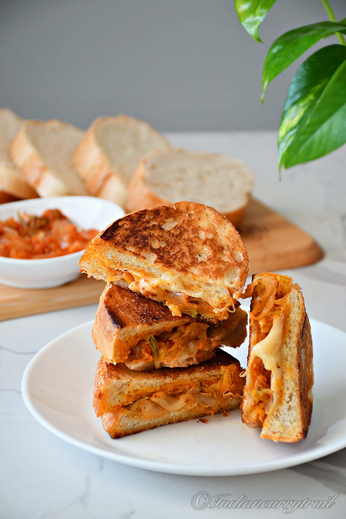 a stack of kimichi grilled cheese sandwich kept on a plate and a bowl of kimichi and breadslices behind