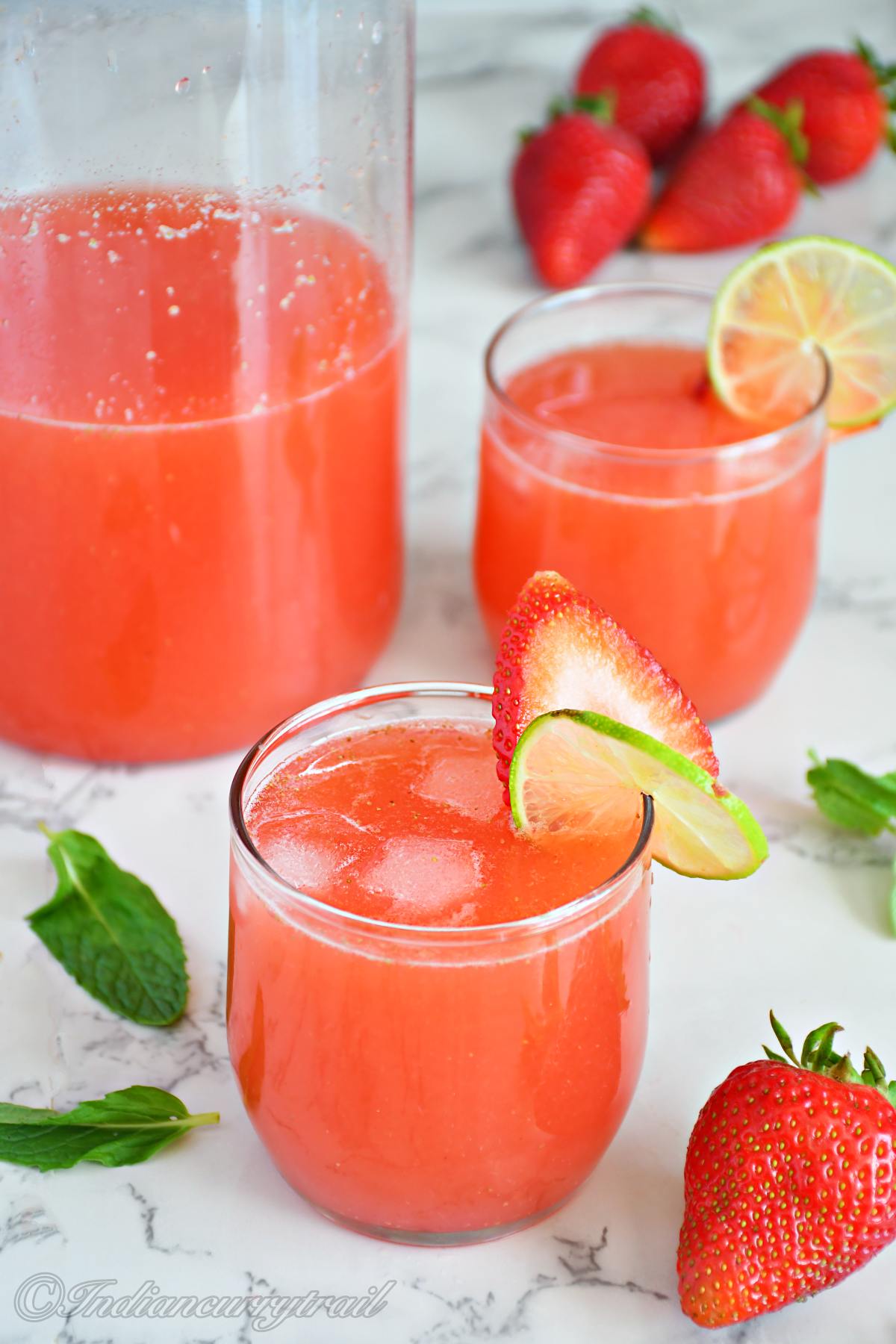 a glass of strawberry agua fresca decorated with a slice of lime and strawberry kept near another glass and a pitcher which has strawberry agua fresca