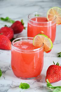 closeup shot showing a glass of strawberry agua fresca decorated with a slice of lime and strawberry kept near another glass and strawberries and mint leaves on the floor