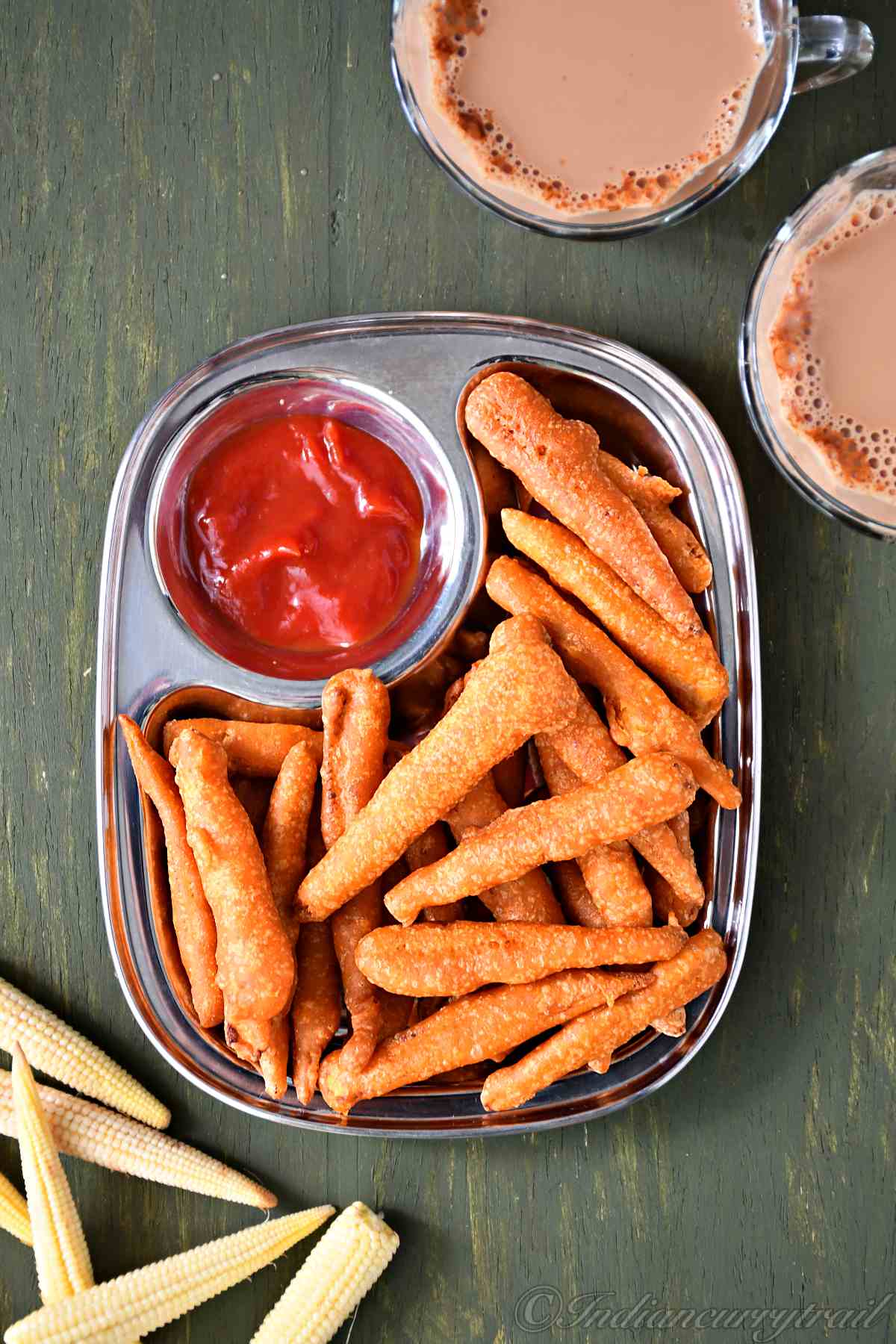 top view of baby corn golden fry, arranged on a plate with ketchup on the side and a couple of tea cups and raw baby corn kept near to it