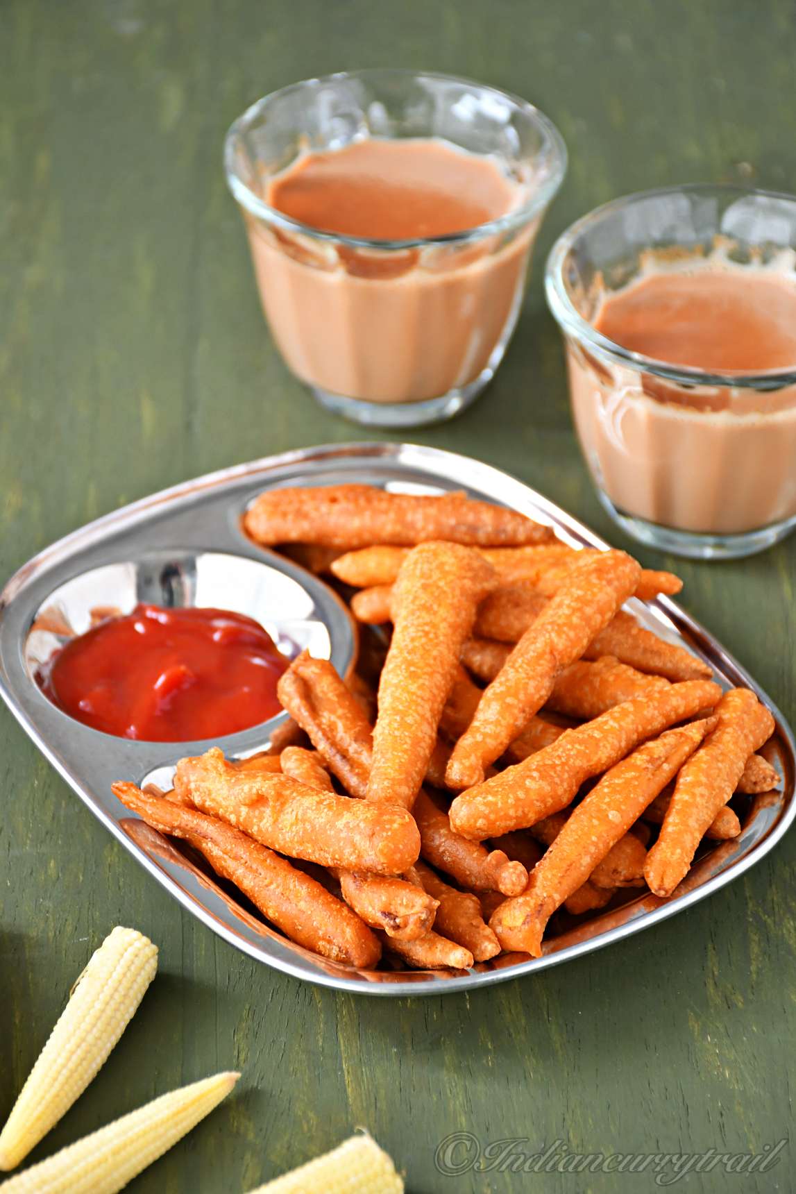 a small plate filled with deep-fried golden baby corn with tomato ketchup and a couple of teacups and raw baby corn kept nearby